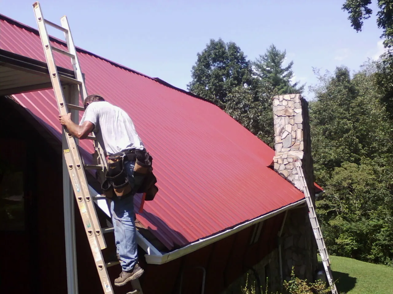 Roofer on a ladder beside a red metal roof and stone chimney — Soffit & Fascia Repair by Roofing Brothers in Hermitage